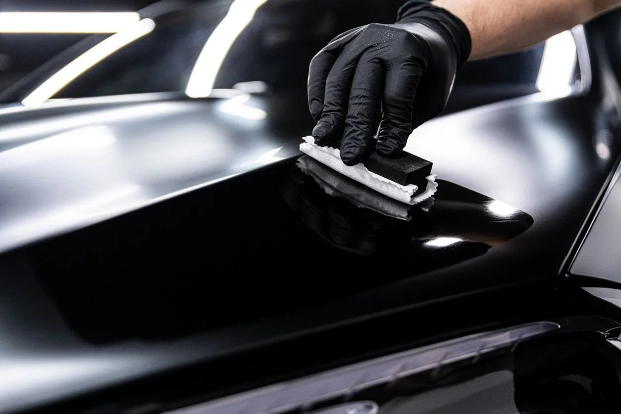 A person wearing black gloves polishing the surface of a black car with a polishing pad and a cloth.