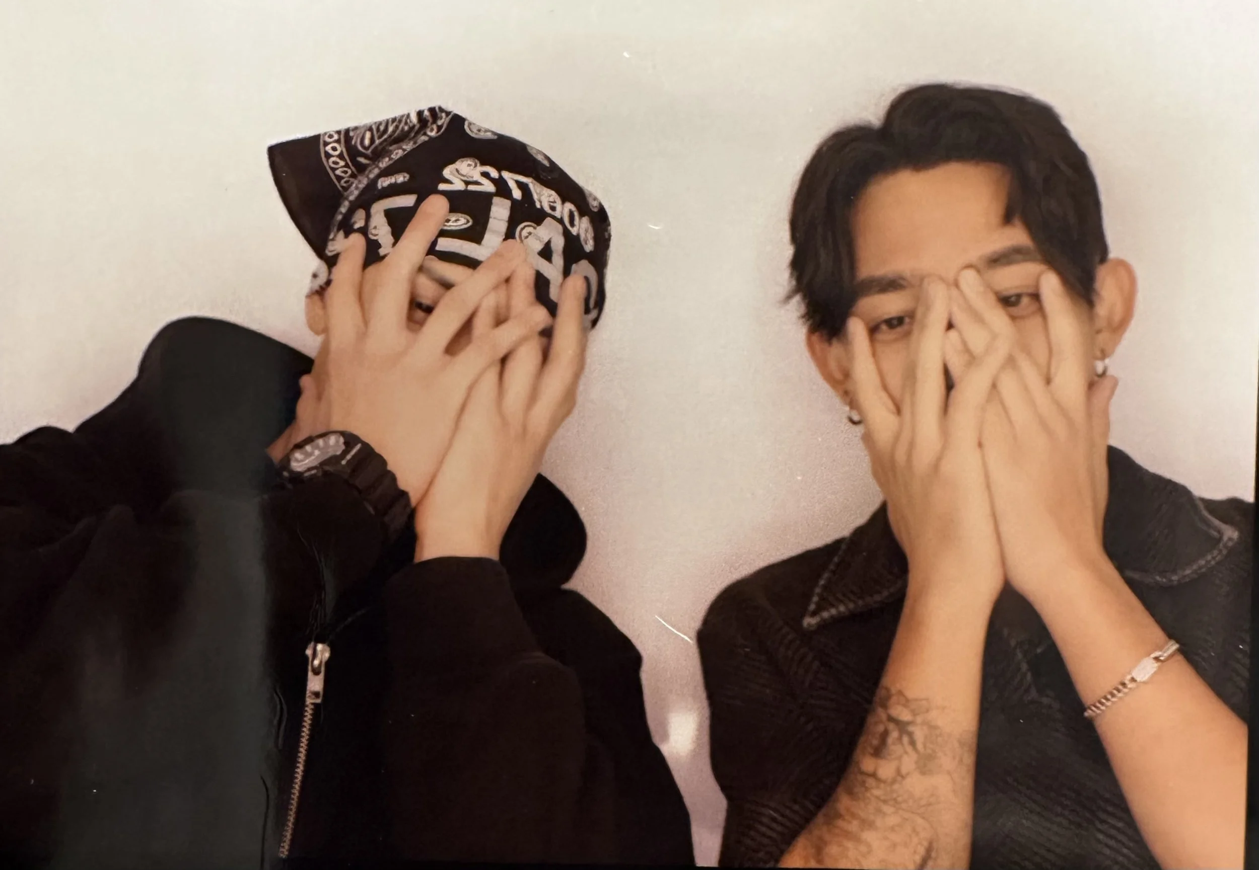 Two people covering their faces with their hands, one wearing a bandana and the other with glasses, against a plain background.