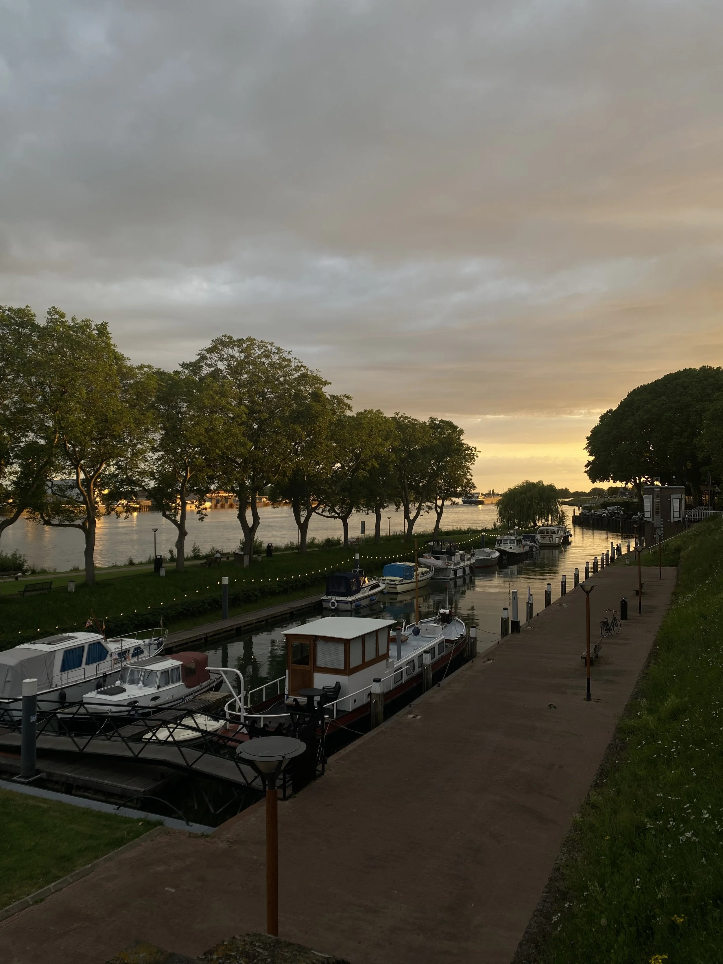 Het uitzicht van "Het Poort Juweel" aan de Lek met een aanlegsteiger en boten, groene bomen en een wandelpad, onder een bewolkte hemel tijdens zonsondergang.