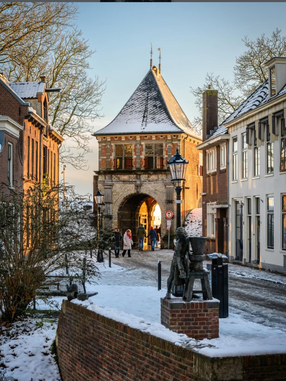 Historisch poortgebouw met stenen en bakstenen muren, een toren met een steil hellend dak bedekt met sneeuw, omringd door oude gebouwen, enkele mensen wandelen er doorheen, in de voorgrond een beeldhouwwerk van een mens met een pot" Het Poort Juweel