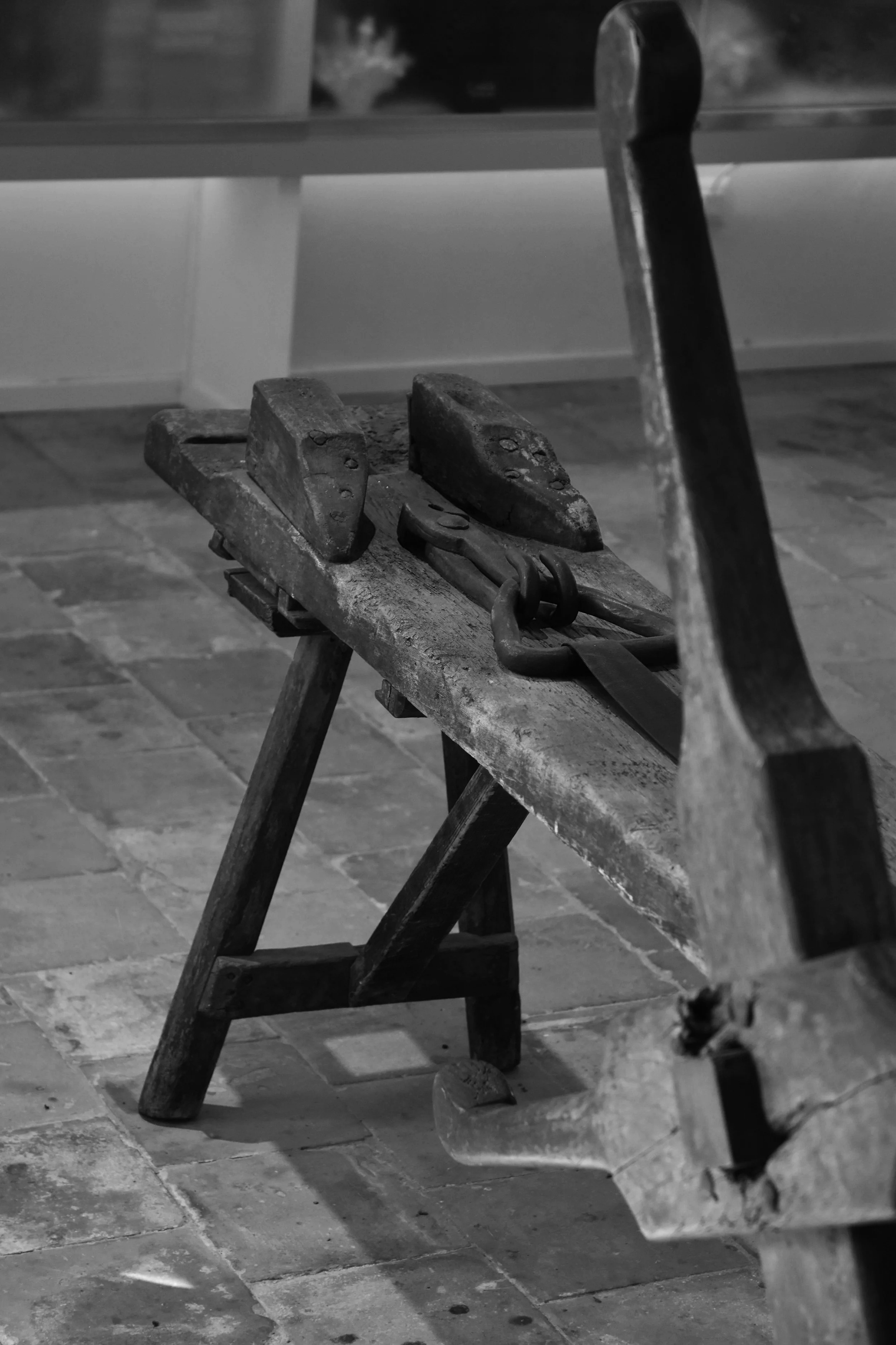 Een oude, houten werkbank met gereedschap zoals vleestangen en een koevoet, staand op een stenen vloer in een werkruimte.