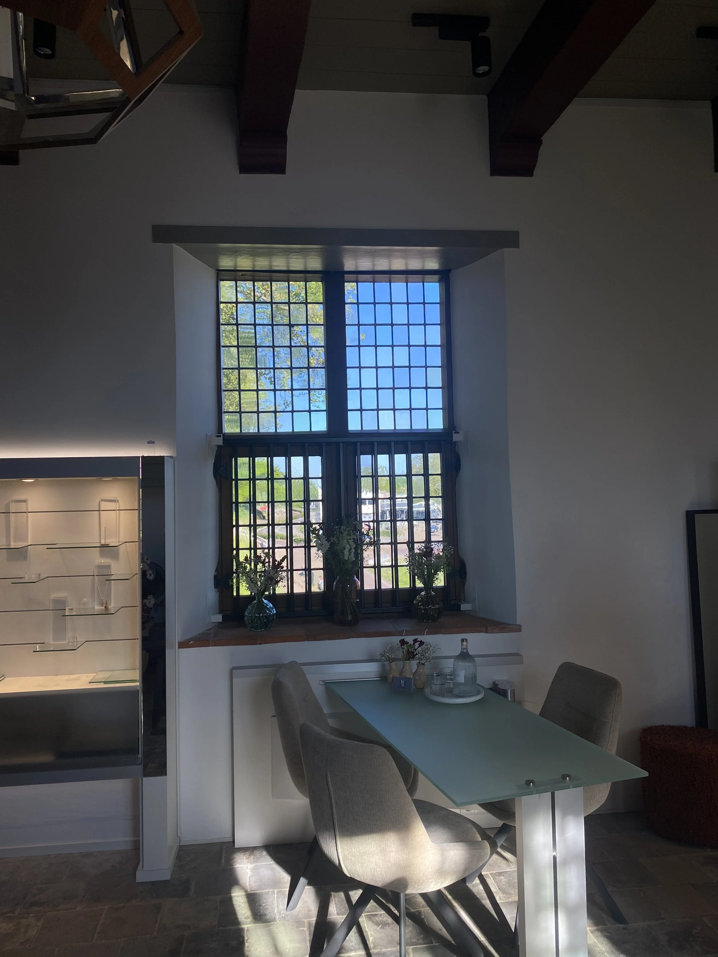 Binnenaanzicht van de winkel van Edelsmederij Puck Huizer in het porotjuweel. Hier worden opdrachten ontworpen een kamer met grote raam waarop bomen en blauwe lucht zichtbaar zijn, een tafel met vazen en bloemen, en twee gestoffeerde stoelen.