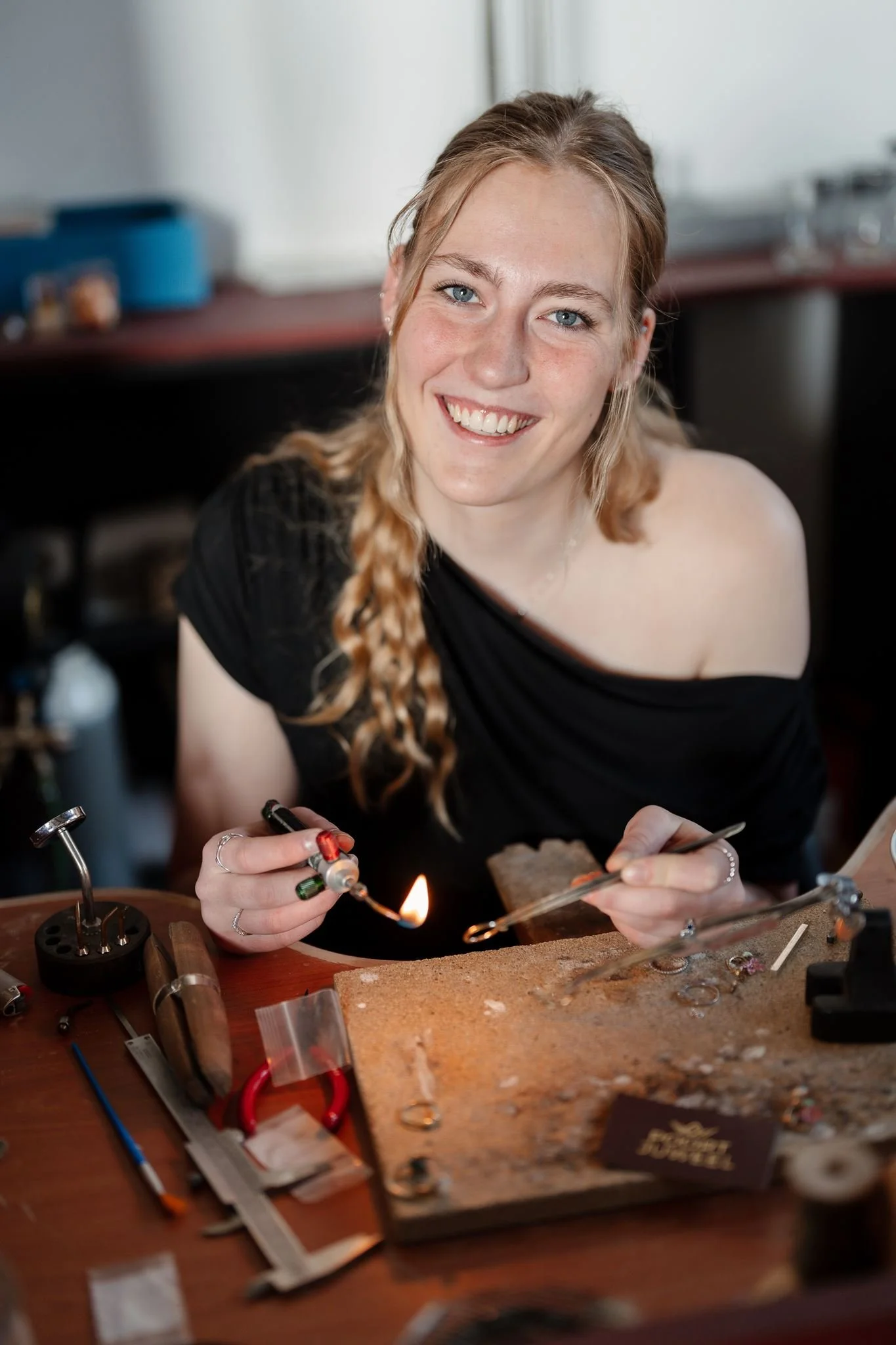 Een jonge vrouw met lang, blond, gekruld haar en blauwe ogen werkt aan het maken van sieraden bij een werkbank, met verschillende gereedschappen en materialen rondom haar.