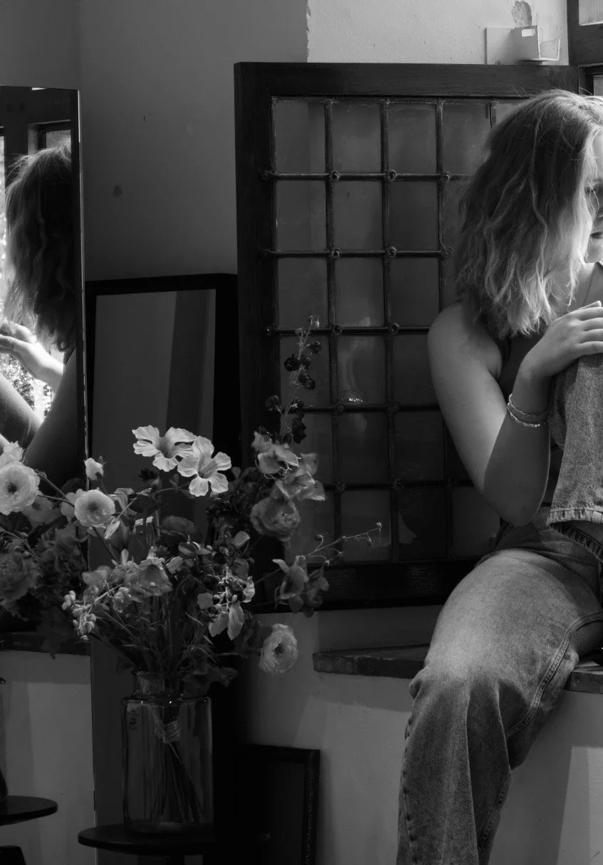 Een zwart-wit foto van een vrouw met golvend haar die op een vensterbank zit, haar arm om een persoon heen waarvan alleen de schouder en een deel van het gezicht zichtbaar zijn. Op de voorgrond staat een vaas met bloemen, en er is een spiegel en een houten kamerscherm op de achtergrond.