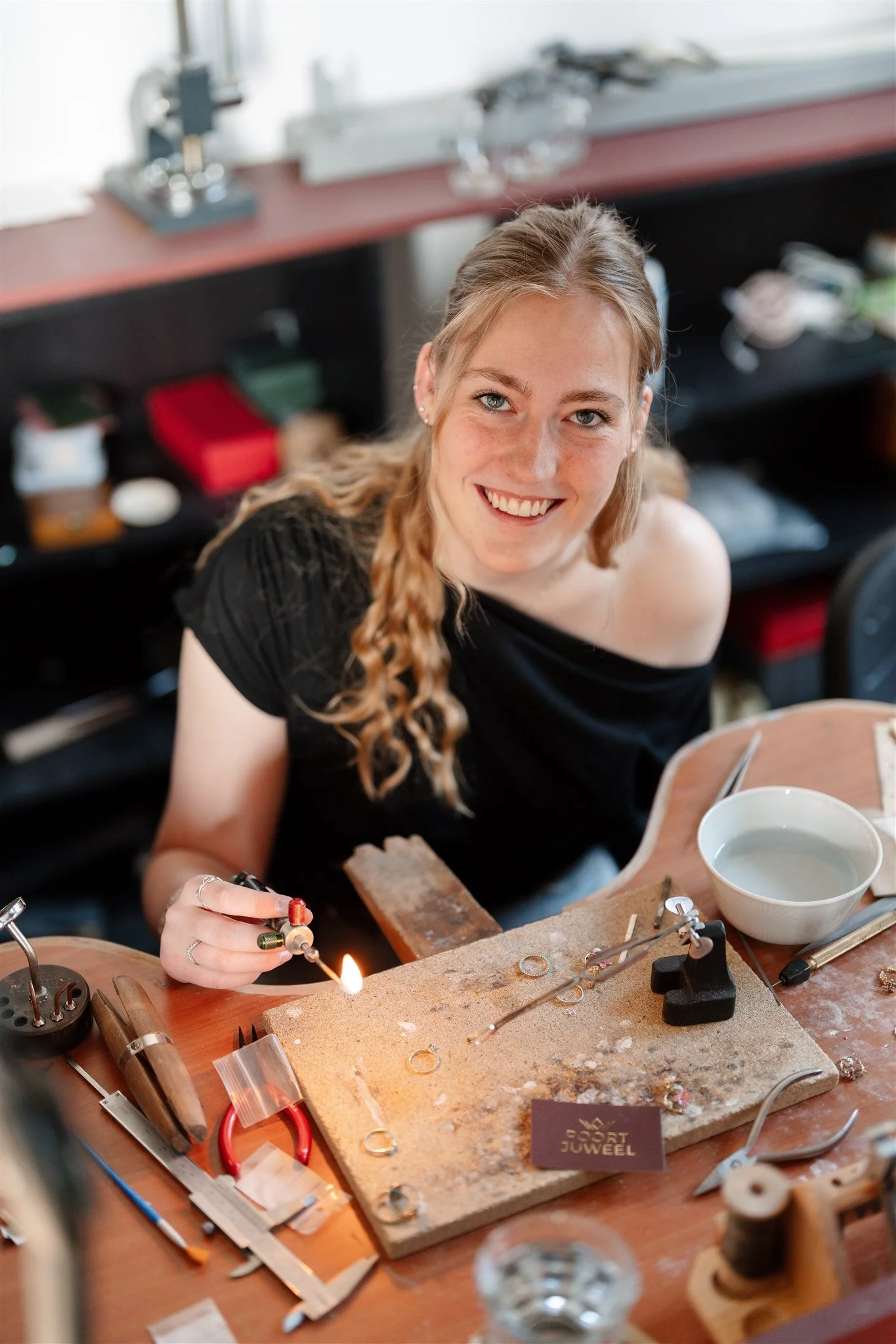Een vrouw aan een werktafel met juweliersgereedschap, die een ring samenstelt met een vlam van een soldeerblam, omringd door gereedschap en materialen op de werkbank.