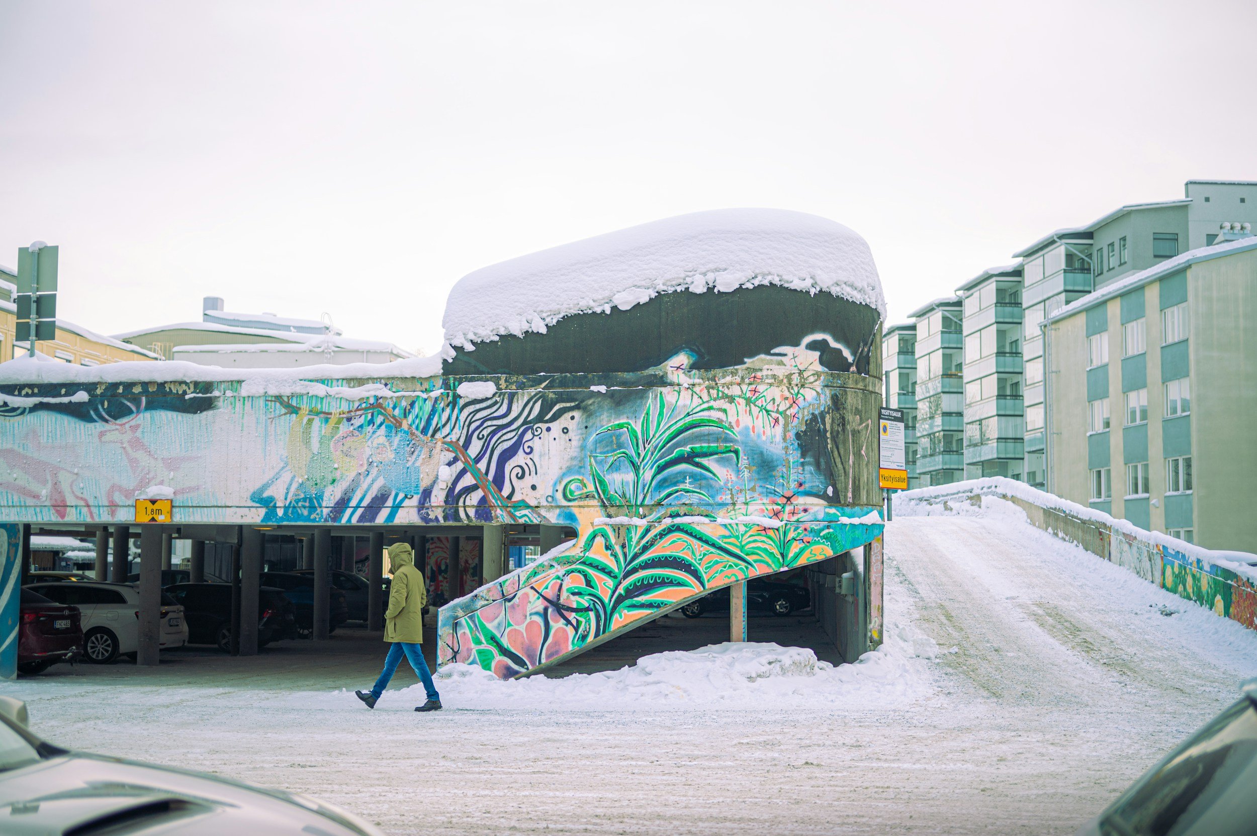 Snow-covered urban graffiti structure with a colorful mural, metal parking garage in the foreground, and residential buildings in the background, during winter.