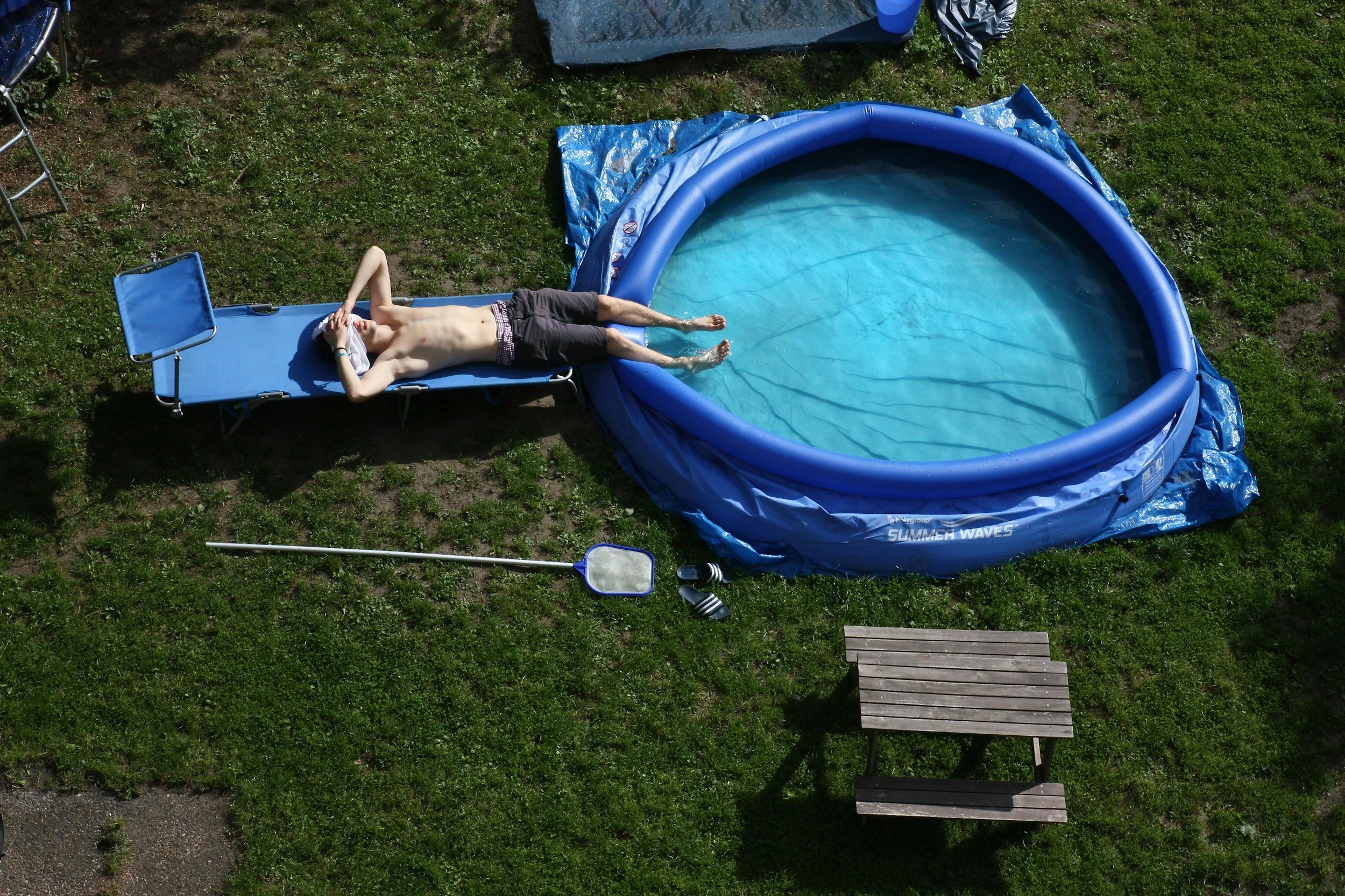 A shirtless man with dark shorts lying on a blue lounge chair next to an inflatable kiddie pool on a grassy yard.