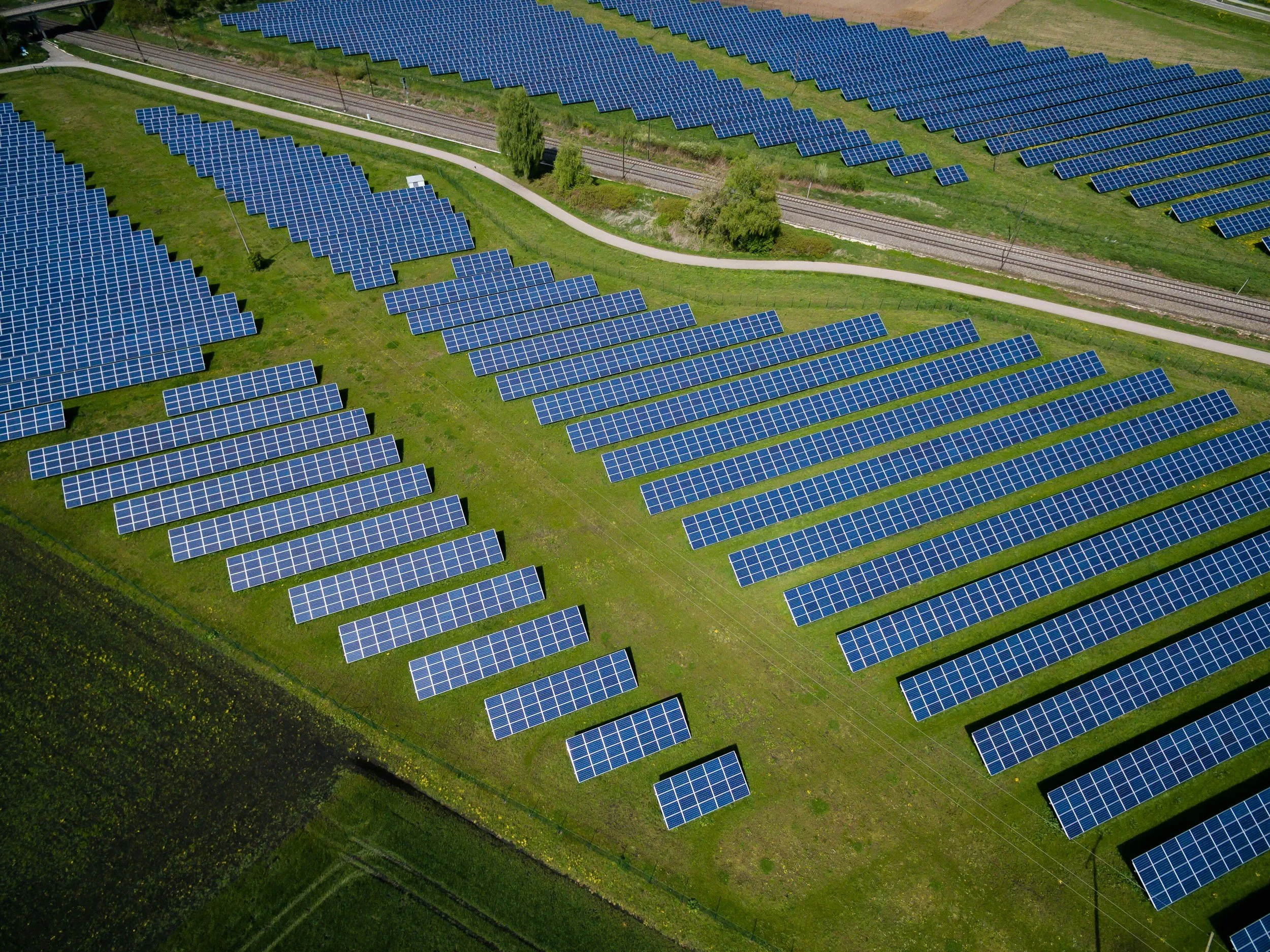 Aerial view of a solar farm with multiple rows of blue solar panels arranged on green grass, divided by a walking path and railway tracks, with trees and open land in the background.