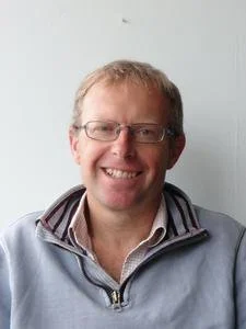 A man with glasses smiling, wearing a light grey jacket and a collared shirt, standing against a plain light background.