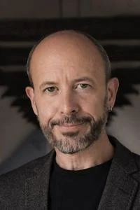 Portrait of a middle-aged man with a beard and mustache, wearing a dark blazer over a black shirt, against a dark background.
