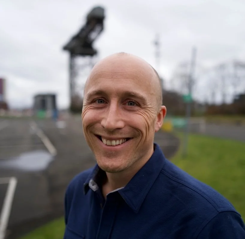 A smiling man with a shaved head in a blue shirt outdoors, with a blurry parking lot and crane in the background.