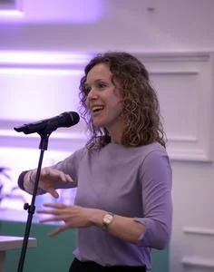 Woman with curly hair speaking into a microphone, smiling and gesturing with her hands in a well-lit room.
