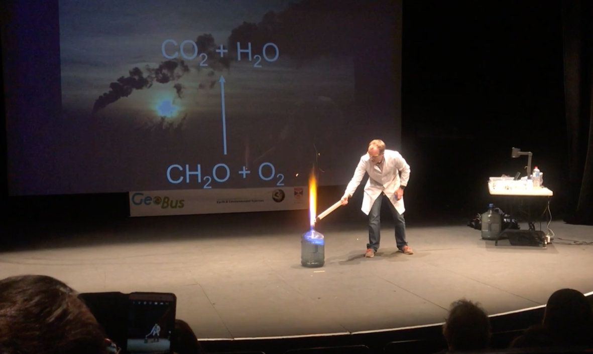 Science demonstration showing a chemical reaction involving an open container with a flame on top, a person in a white lab coat tending to it, a science presentation slide with chemical formulas, and an audience observing.