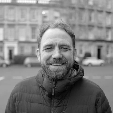 A smiling man with a beard wearing a zipped-up jacket outdoors, with a city street and buildings in the background.