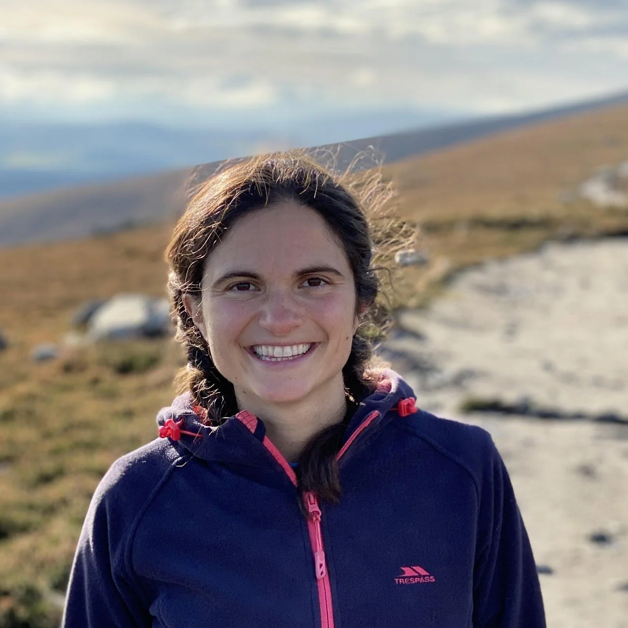 A young woman with dark brown hair in braided pigtails, smiling outdoors on a cloudy day, wearing a navy blue jacket with pink accents, standing on a trail in a grassy hilly area.