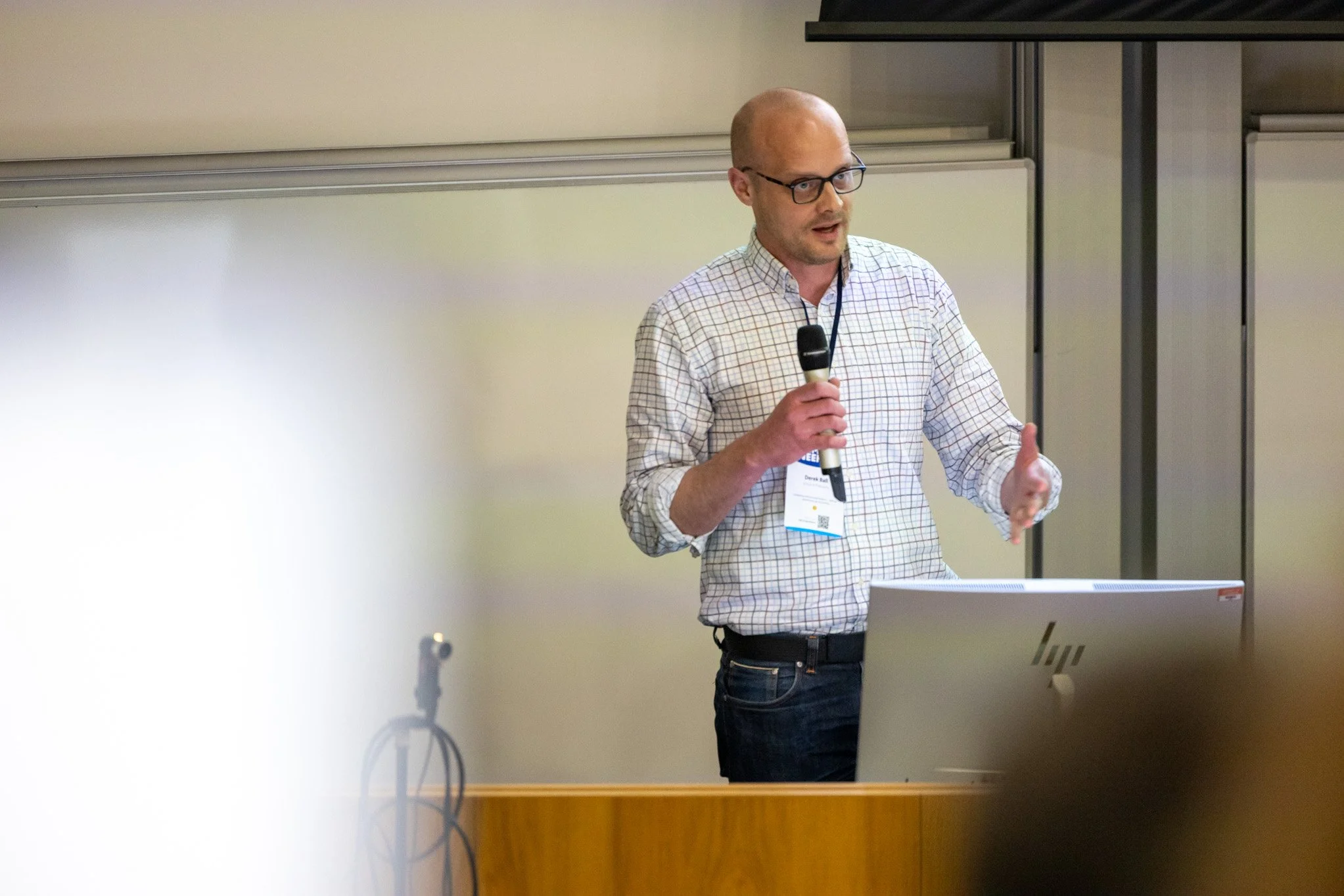 A man with glasses and a checkered shirt giving a presentation in front of a whiteboard, holding a microphone.
