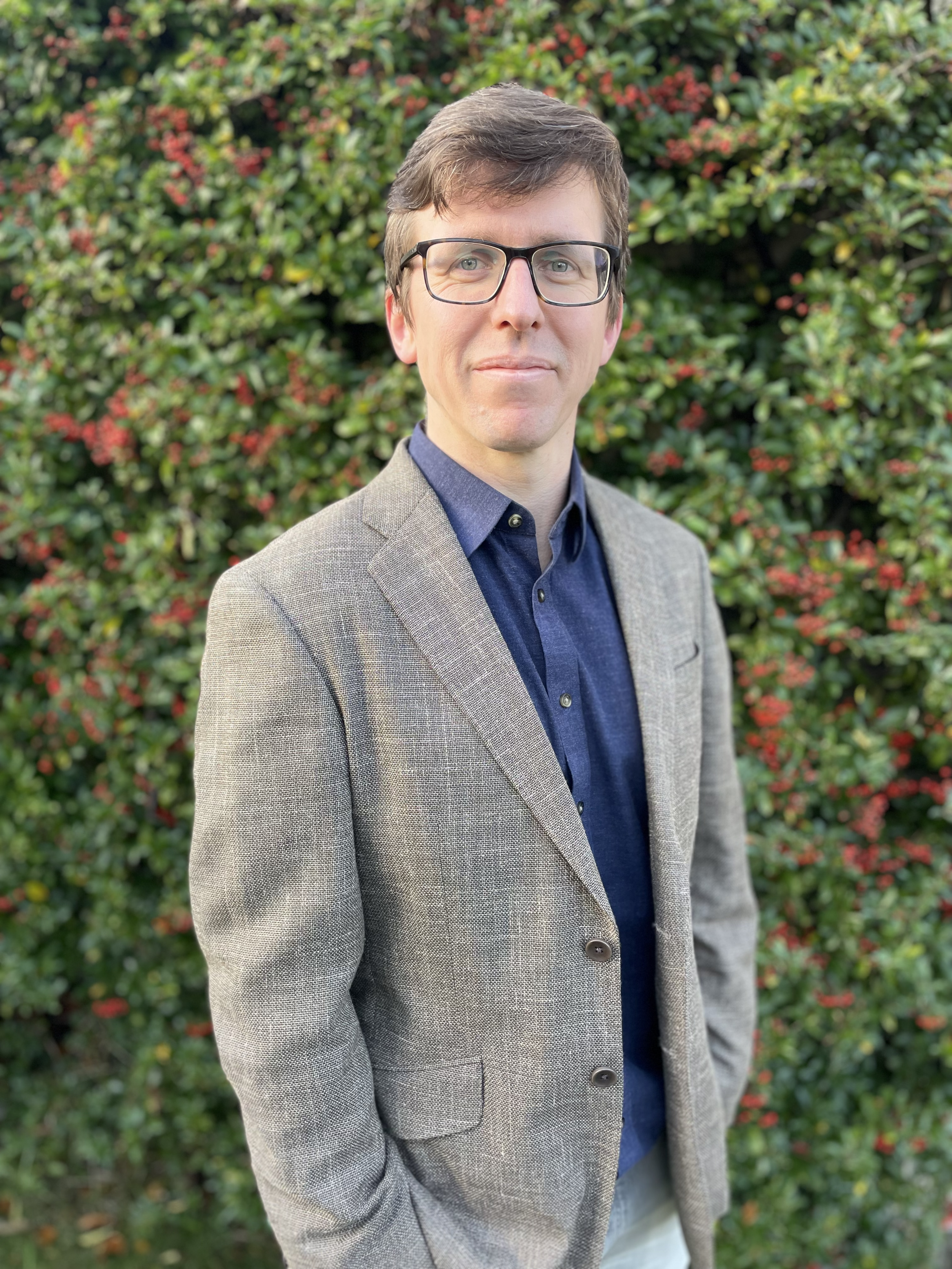 A man with brown hair, glasses, wearing a gray blazer and a blue shirt, standing outdoors in front of a dense hedge with red and green leaves.
