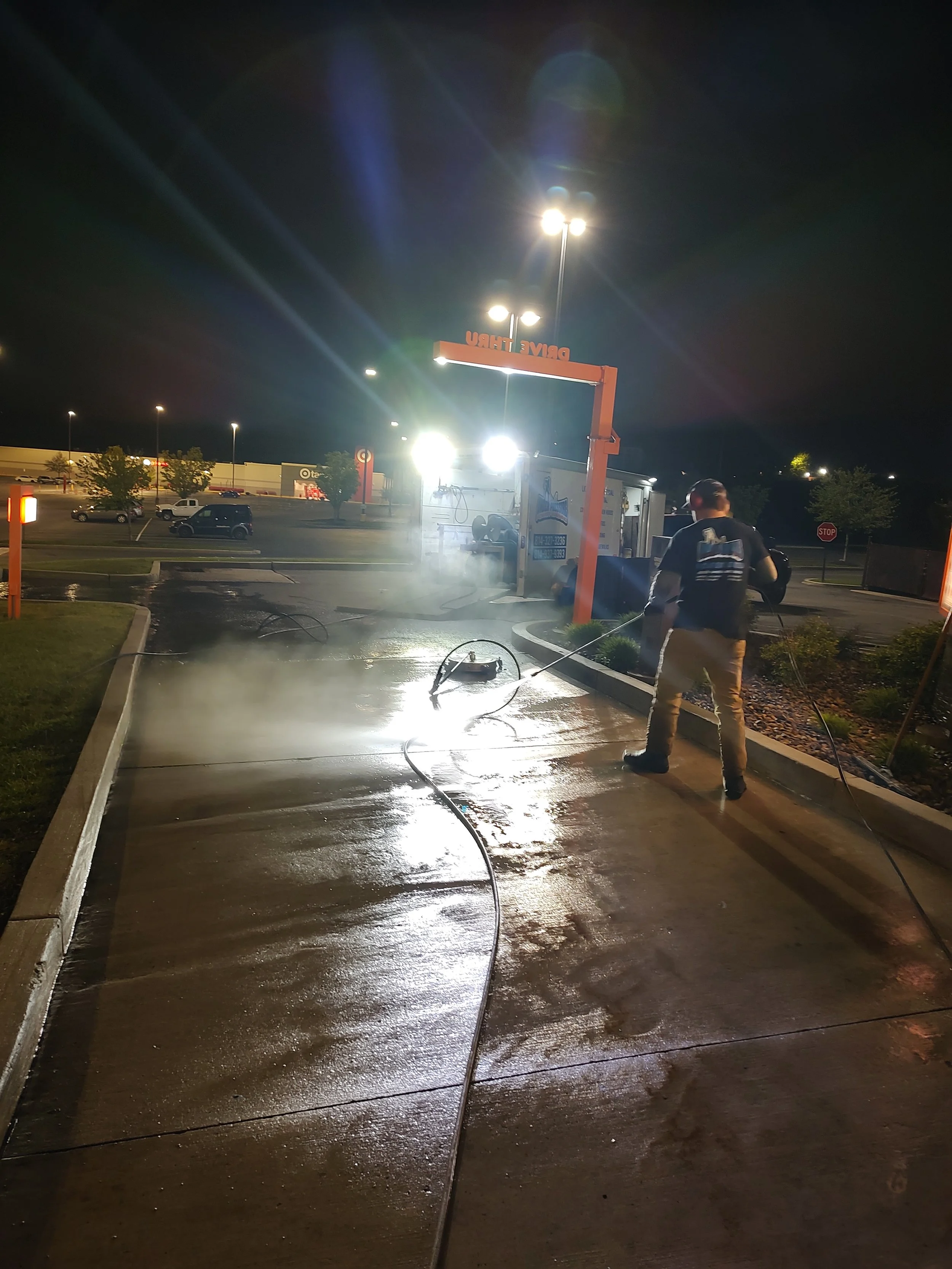 A person cleaning a concrete sidewalk at night with high-pressure water, with bright lights illuminating the scene, parking lots, trees, and a store visible in the background.
