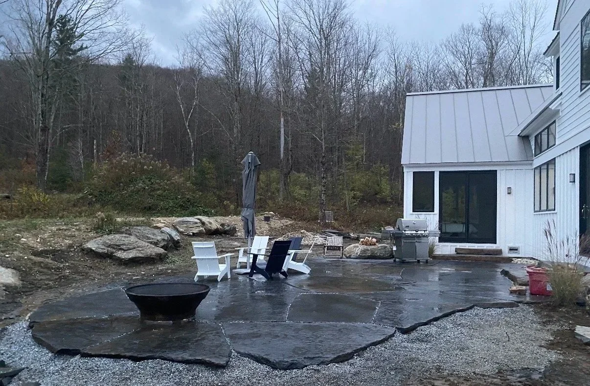 A backyard patio with large black stone slabs, white and black chairs, a closed patio umbrella, a grill, and a fire pit, with a white house on the right and a wooded hill in the background.