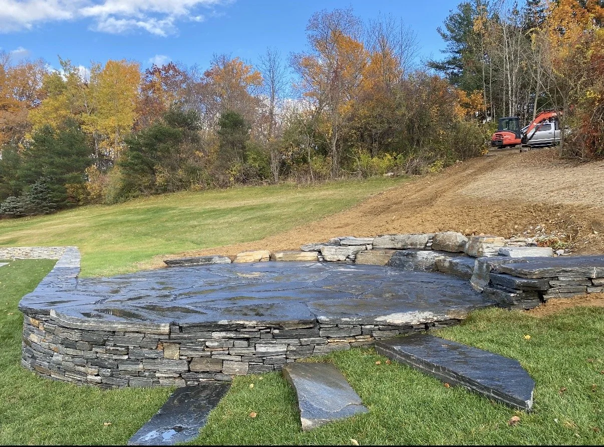 A newly constructed stone patio with built-in steps, located on a grassy lawn, with trees showing autumn foliage in the background. There is a hill with a construction vehicle parked at the top right corner.