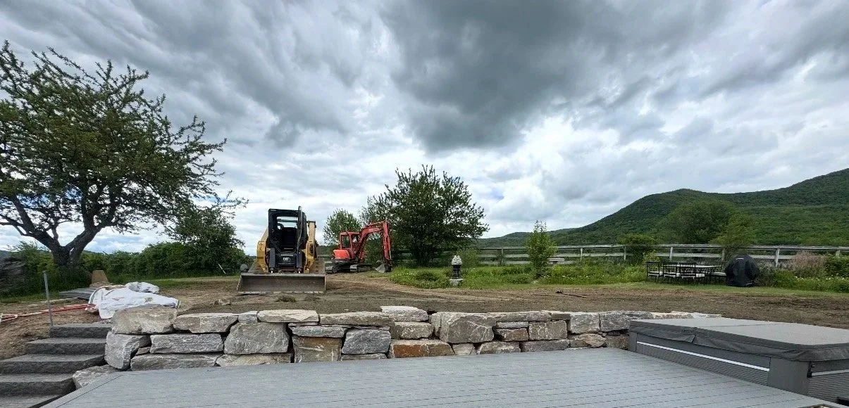 Construction site with two small excavators, stone wall, and outdoor scenery including trees, hills, cloudy sky, picnic table, and a hot tub cover.