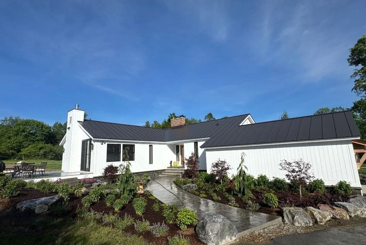 White house with black metal roof, surrounded by a well-maintained garden with various plants and rocks, under a blue sky