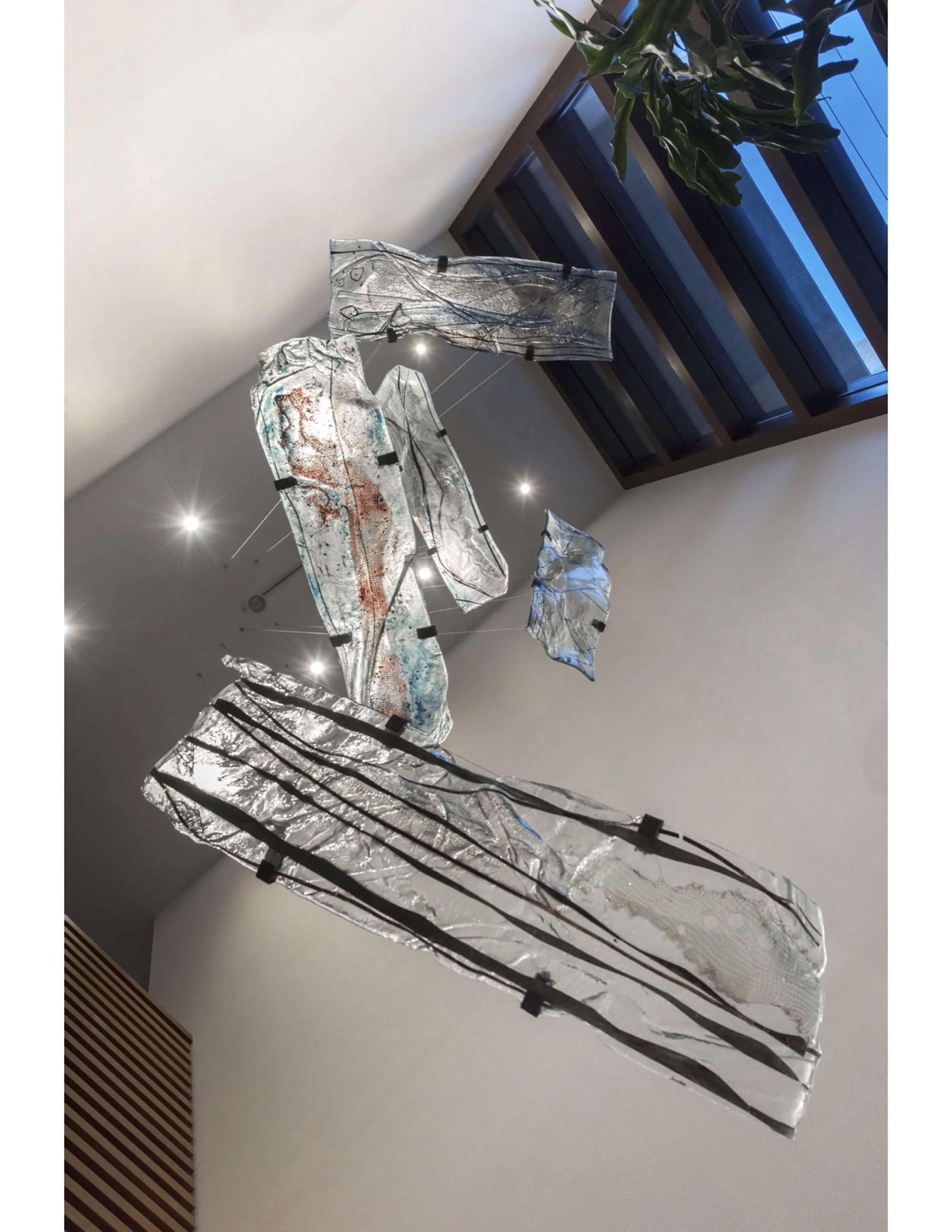 A suspended aerial installation of five curved, irregular glass pieces acting as a family portrait. Hanging horizontally in an 8-meter foyer next to a large window, the organically shaped elements capture natural light to reveal permanent color gradi