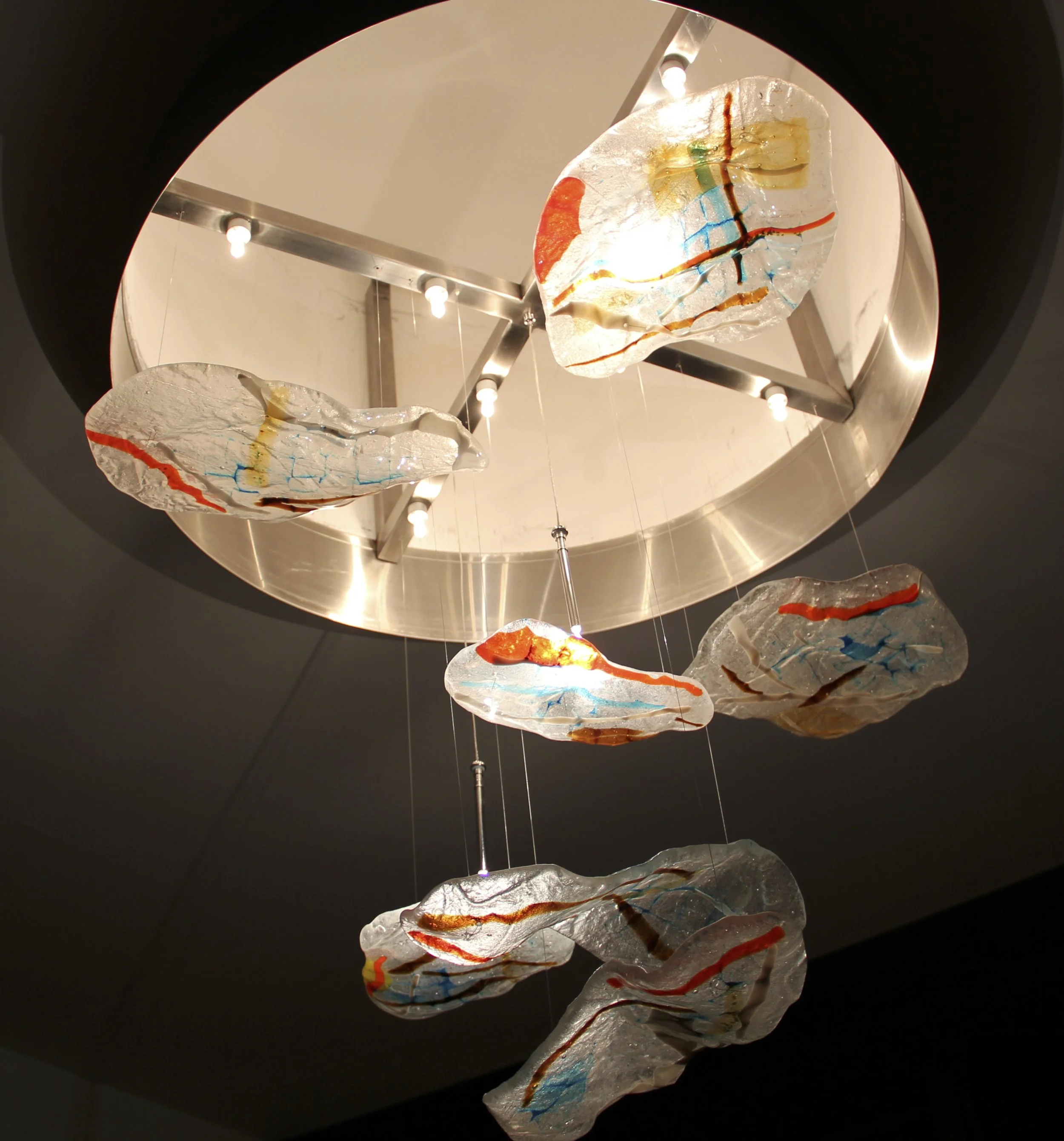 A custom contemporary chandelier designed to make thick glass appear weightless. The curved crystal canvases catch pinpoint light from above to illuminate their ice-like textures and internal flows of red, blue, and amber glass.
