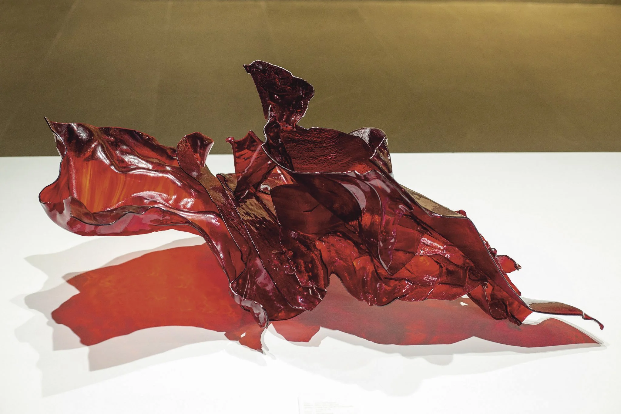 A structural exploration of chromatic depth where light reveals hidden internal hues of red, yellow and gold within the glass. The fluid, curved composition extends into the surrounding space by casting a vivid red shadow onto the surface below.
