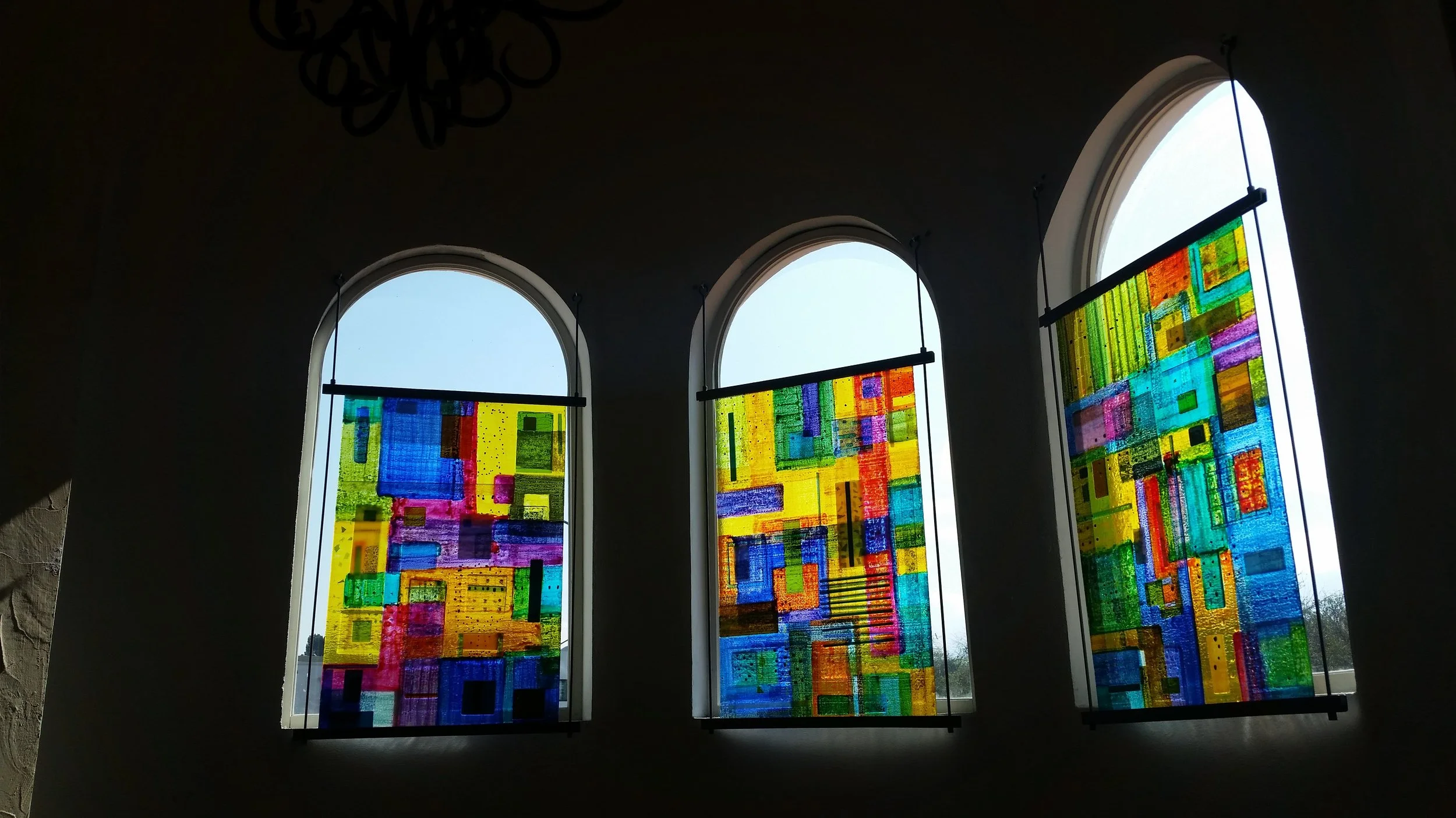 A contemporary three-panel architectural glass installation designed for a residential space. Suspended within arched windows, the glass canvases blend translucent and opaque color fields that filter changing natural light, transforming the interior 