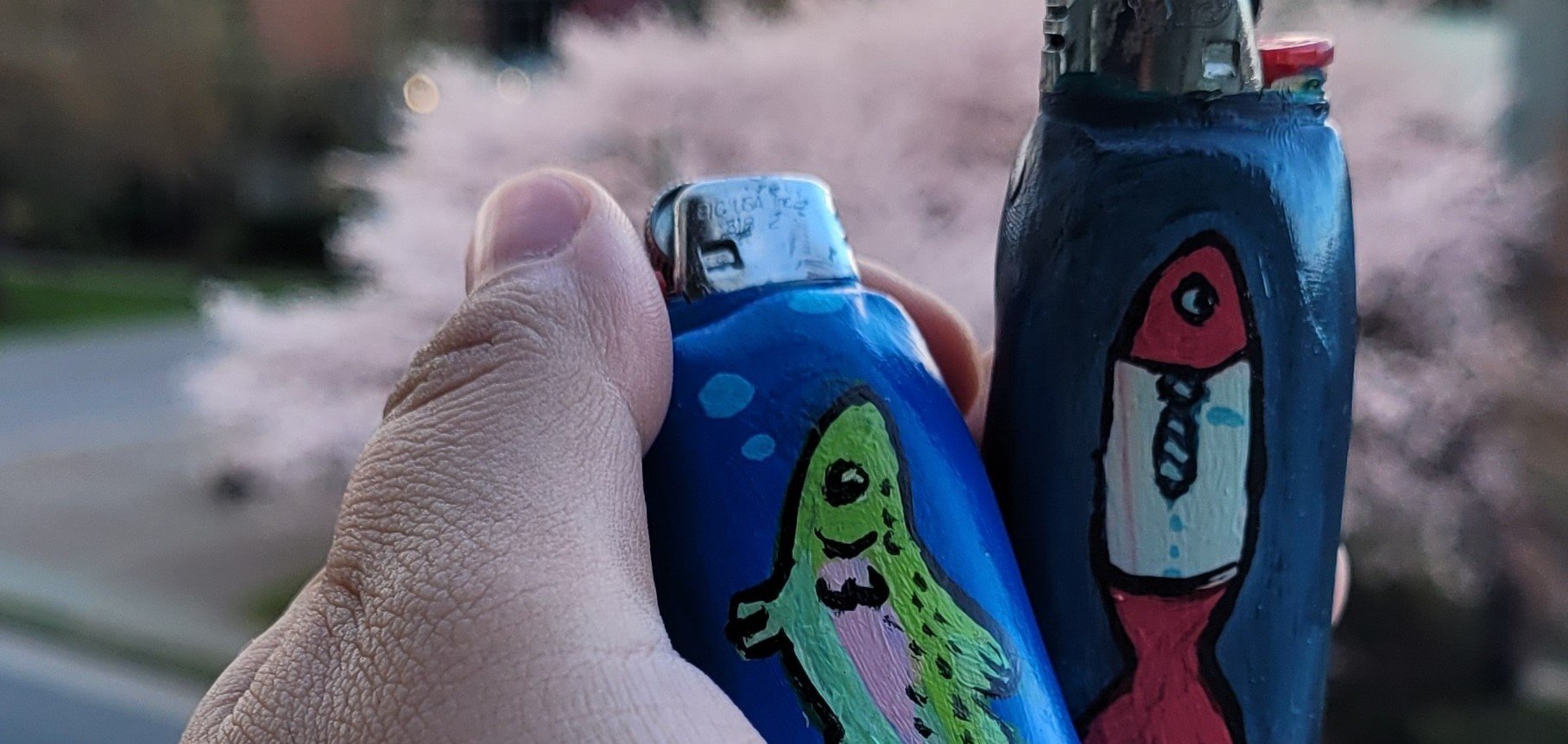 A hand holding two small painted bottles with cartoonish designs, one featuring a green dinosaur and the other showing a red and white rocket ship with a face, against an outdoor background with blurred foliage.