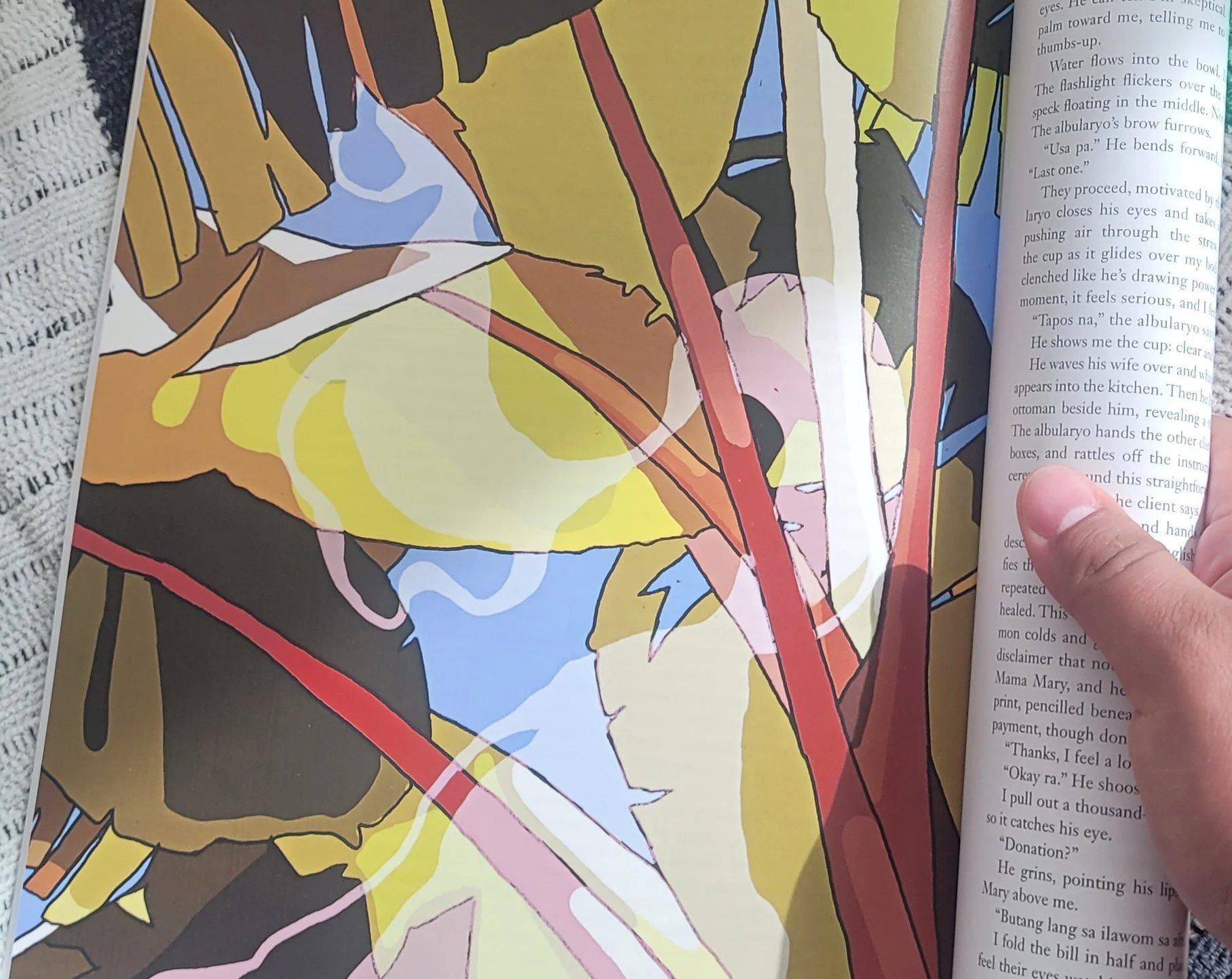 A person holding a book open to a colorful illustration on the left page, with a text passage on the right page. The illustration includes yellow, black, brown, blue, pink, and red colors, depicting a stylized scene with lines and shapes.