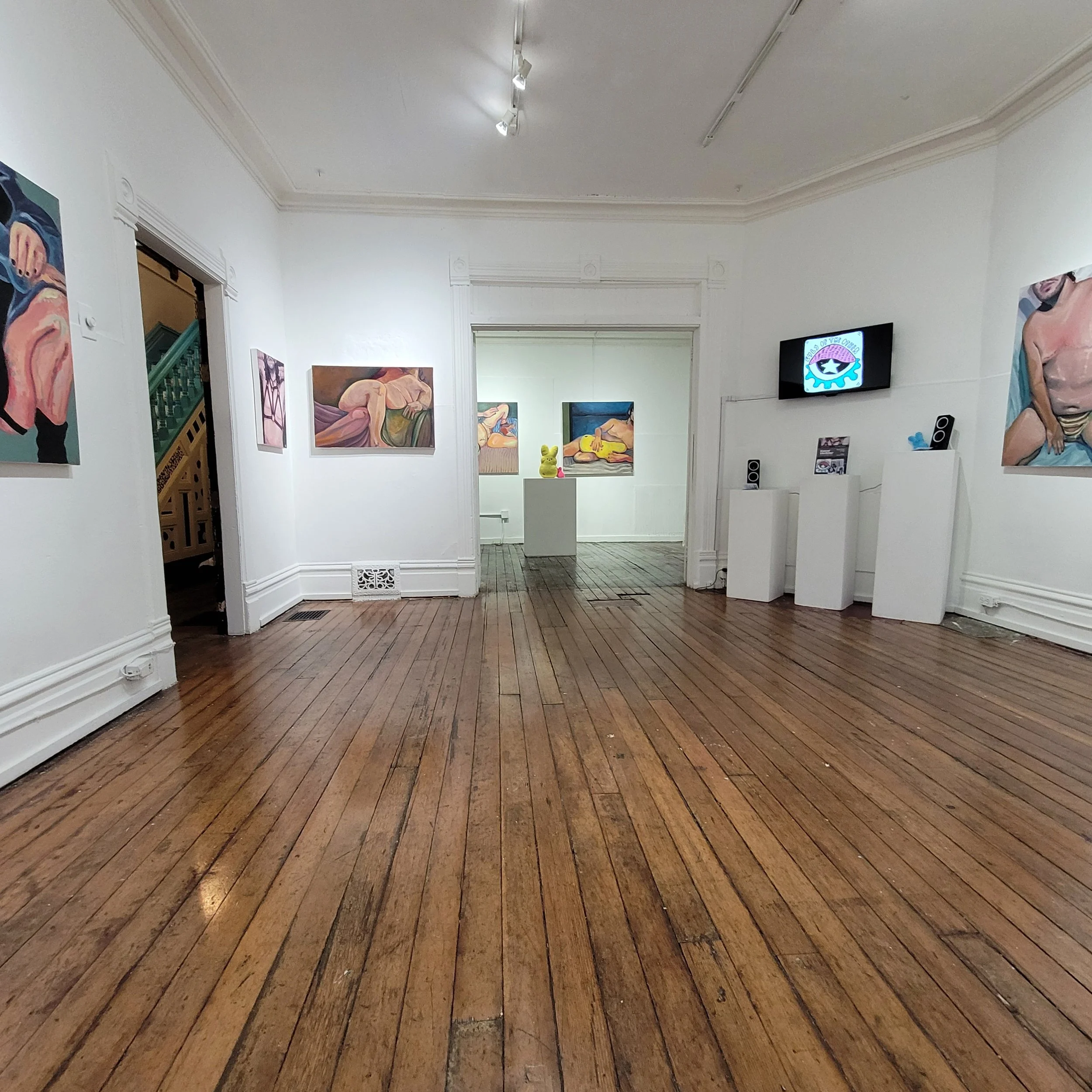 An art gallery with white walls displaying paintings of abstract and figurative artwork, wood flooring, and a mounted television screen showing a logo.