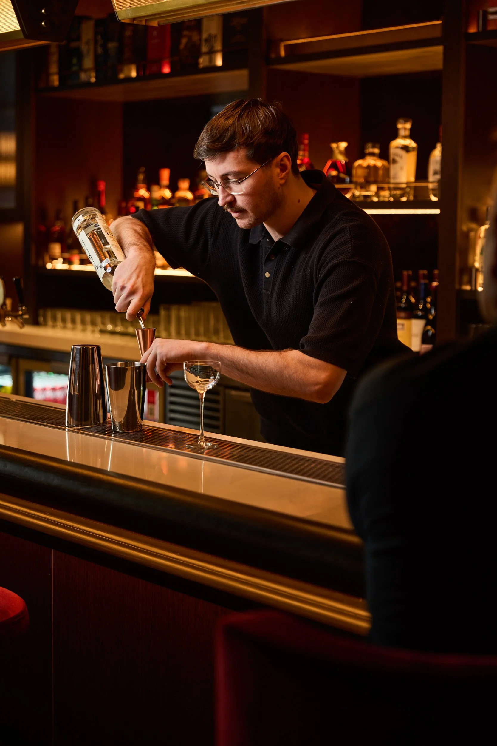 A bartender preparing a cocktail at a bar with shelves of liquor bottles in the background.