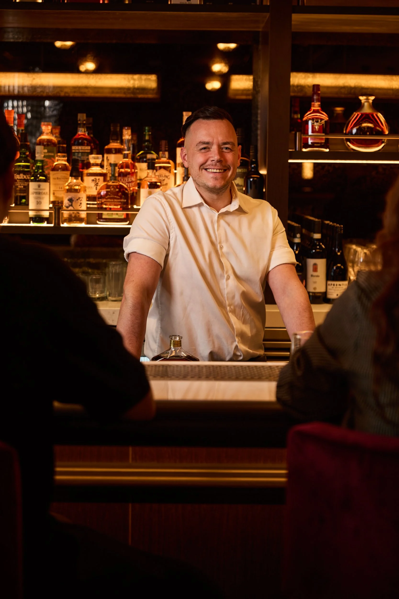 A bartender smiling behind the bar with bottles of liquor in the background, patrons sitting at the bar, dim lighting creating a cozy atmosphere.