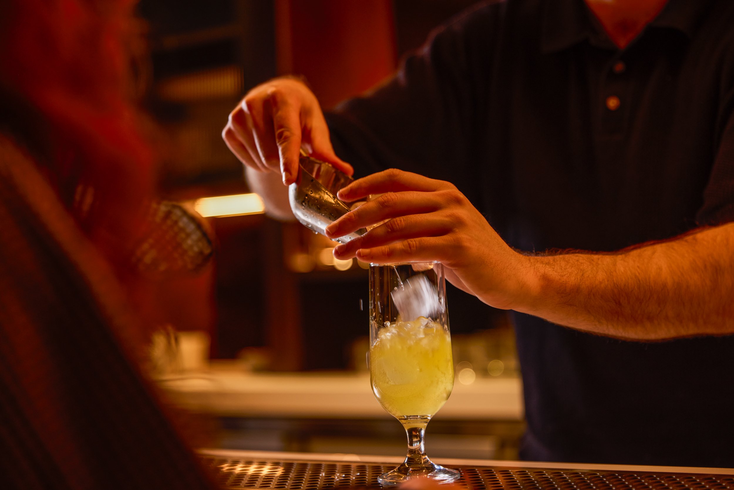 A bartender is preparing a cocktail with ice in a tall glass, using a metal shaker in a dimly lit bar.