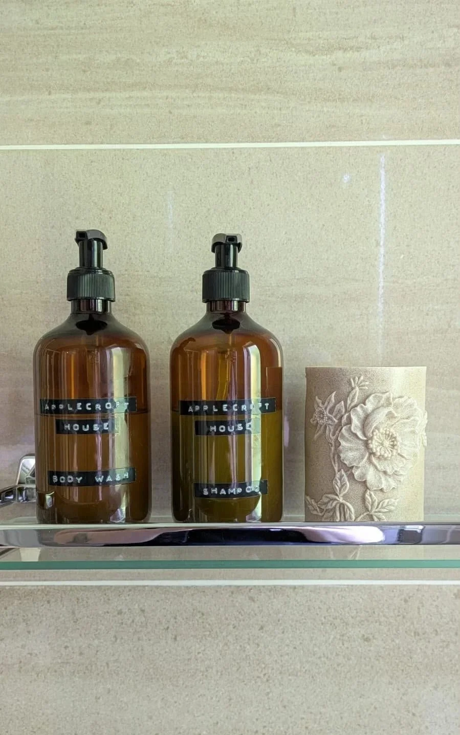 Two amber bottles labeled 'Applecroft House Body Wash' and 'Applecroft House Shampoo' sit next to an embossed floral design on a roll of toilet paper on a glass shelf in a bathroom.