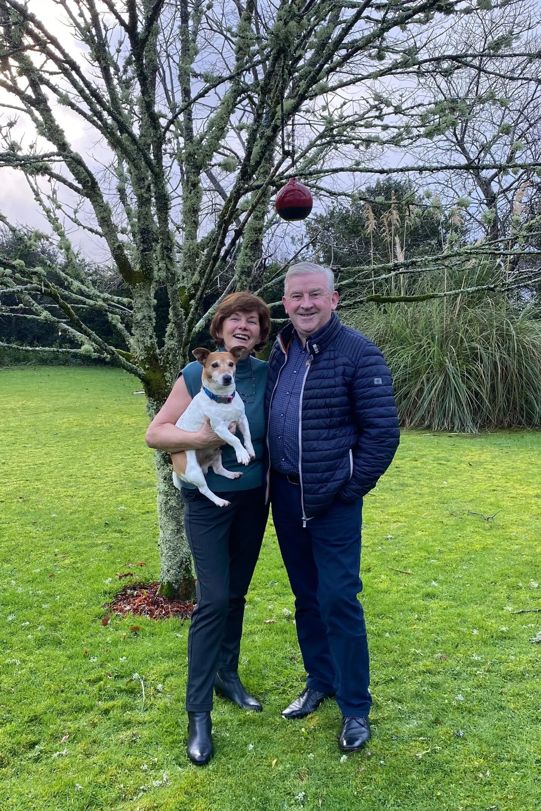 A smiling couple standing on grass in front of a tree, with a small dog and a red Christmas ornament hanging from a branch, during daytime in a park or garden.