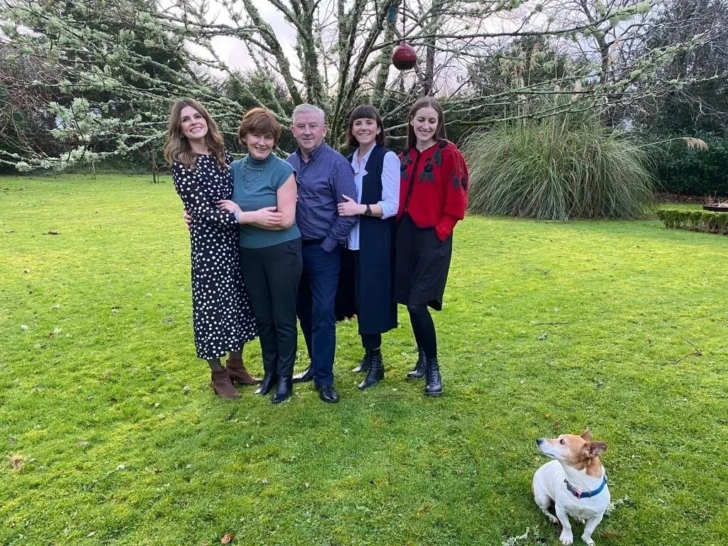 Group of five people standing on a grassy lawn, smiling at the camera, with a dog sitting nearby and a blossoming tree in the background.