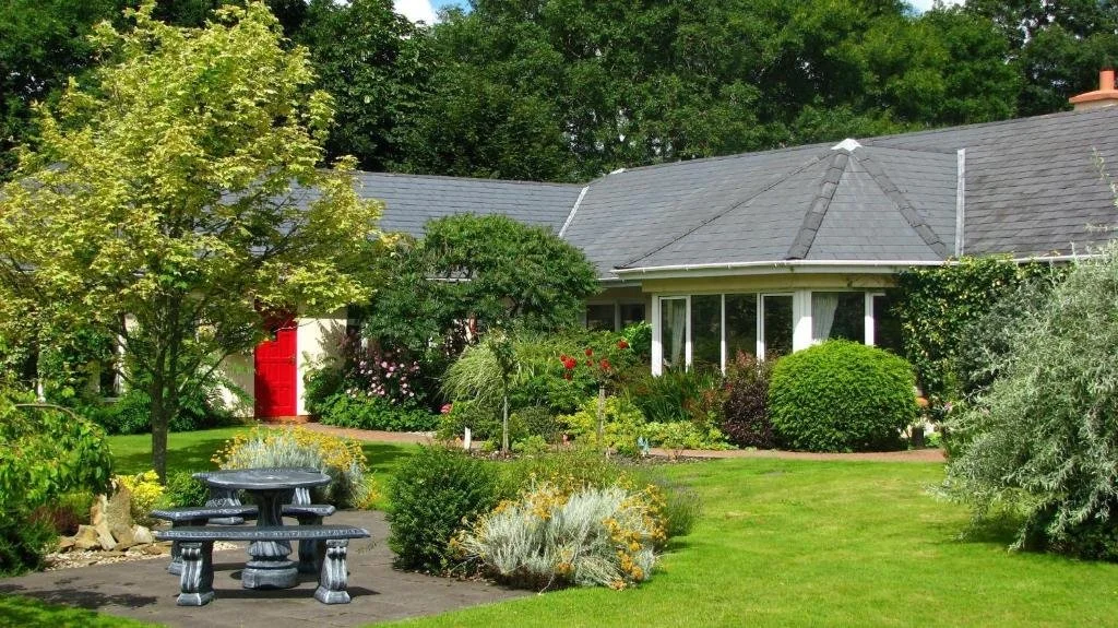 A peaceful backyard garden with a lush green lawn, trees, flowering bushes, and a stone picnic table. There is a house with a gray roof and large windows, a red door, and a variety of plants and shrubs surrounding it.