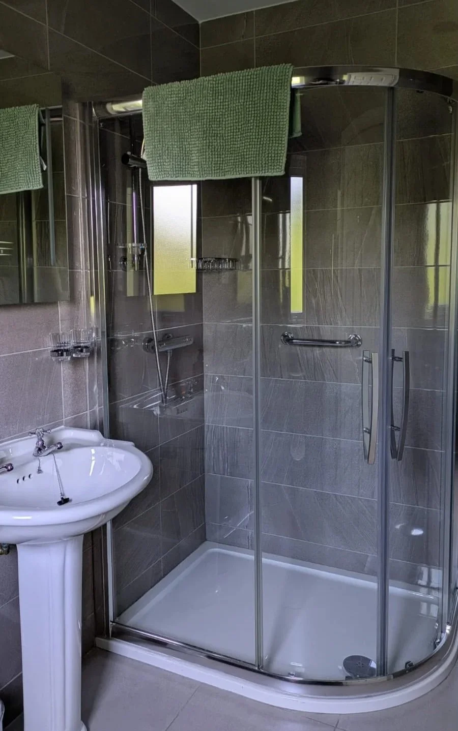 A small bathroom with a corner shower enclosure with glass doors, a white pedestal sink, and green towels hanging on top of the shower. The walls are tiled in dark gray, and there is a frosted window inside the shower.