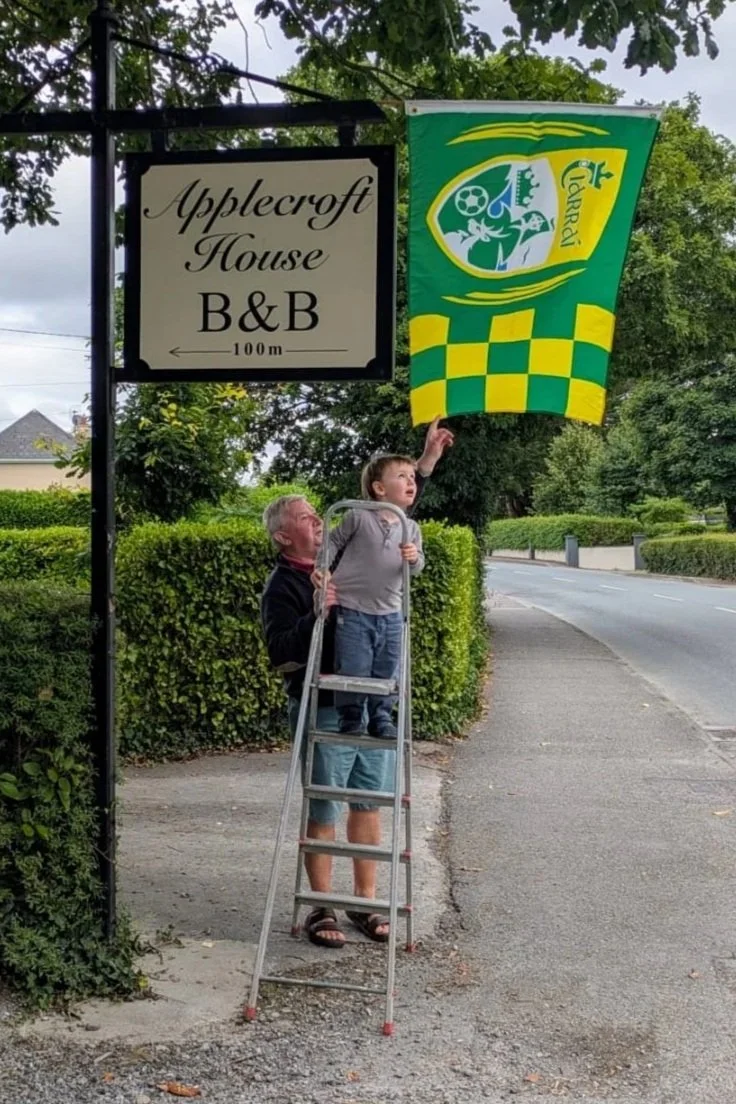 An older man helping a young boy reach a hanging flag on a pole beside a sign that reads 'Applecroft House B&B 100m' along a sidewalk in a neighborhood with greenery and trees.