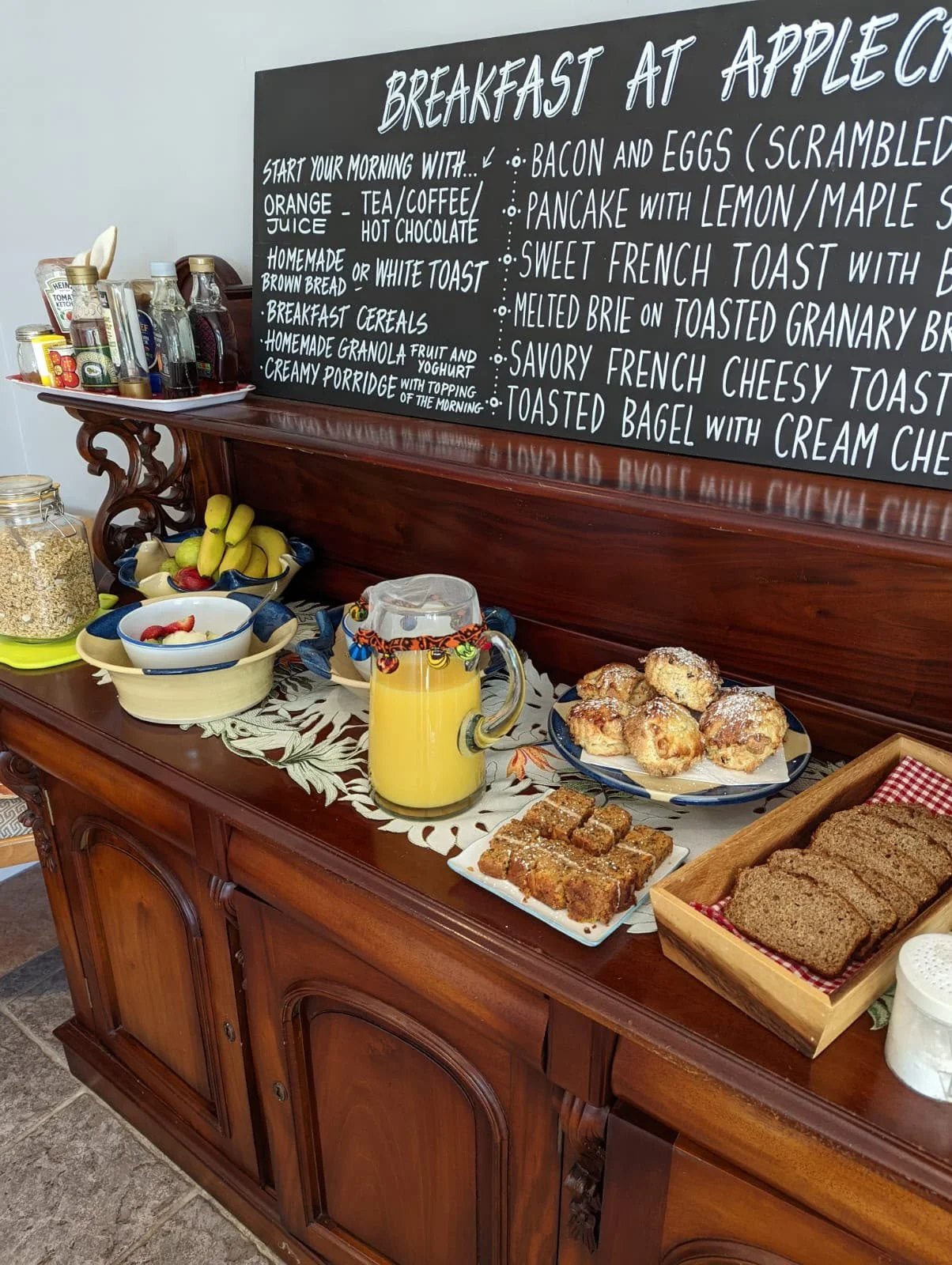 A wooden buffet table with breakfast foods, including muffins, toast, and banana bread, a pitcher of orange juice, a bowl of cereal with strawberries, bananas, and a spoon, and a jar of oats. A large blackboard sign with written breakfast menu items is behind the table, and various condiments and utensils are on a tray to the side.