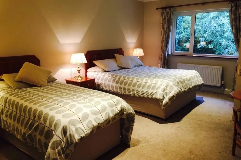 A hotel room with two double beds, nightstands with lamps, a window with floral curtains showing trees outside, and a beige carpet.