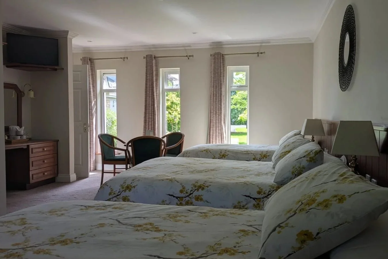 A hotel room with three beds covered in white floral bedding, a small round table with three chairs, a dresser with a mirror, a flat-screen TV in the corner, and three windows with curtains letting in natural light, overlooking greenery outside.