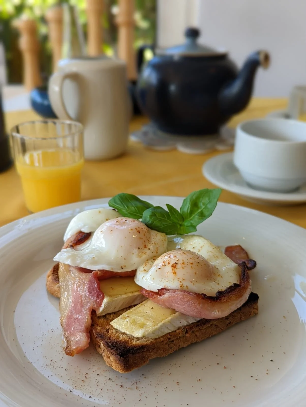 A plate of breakfast featuring toast topped with bacon, eggs, and cheese garnished with basil leaves. In the background, there is a glass of orange juice, a white mug, a black teapot, and a white bowl on a table, with a blurred kitchen or dining area visible behind.