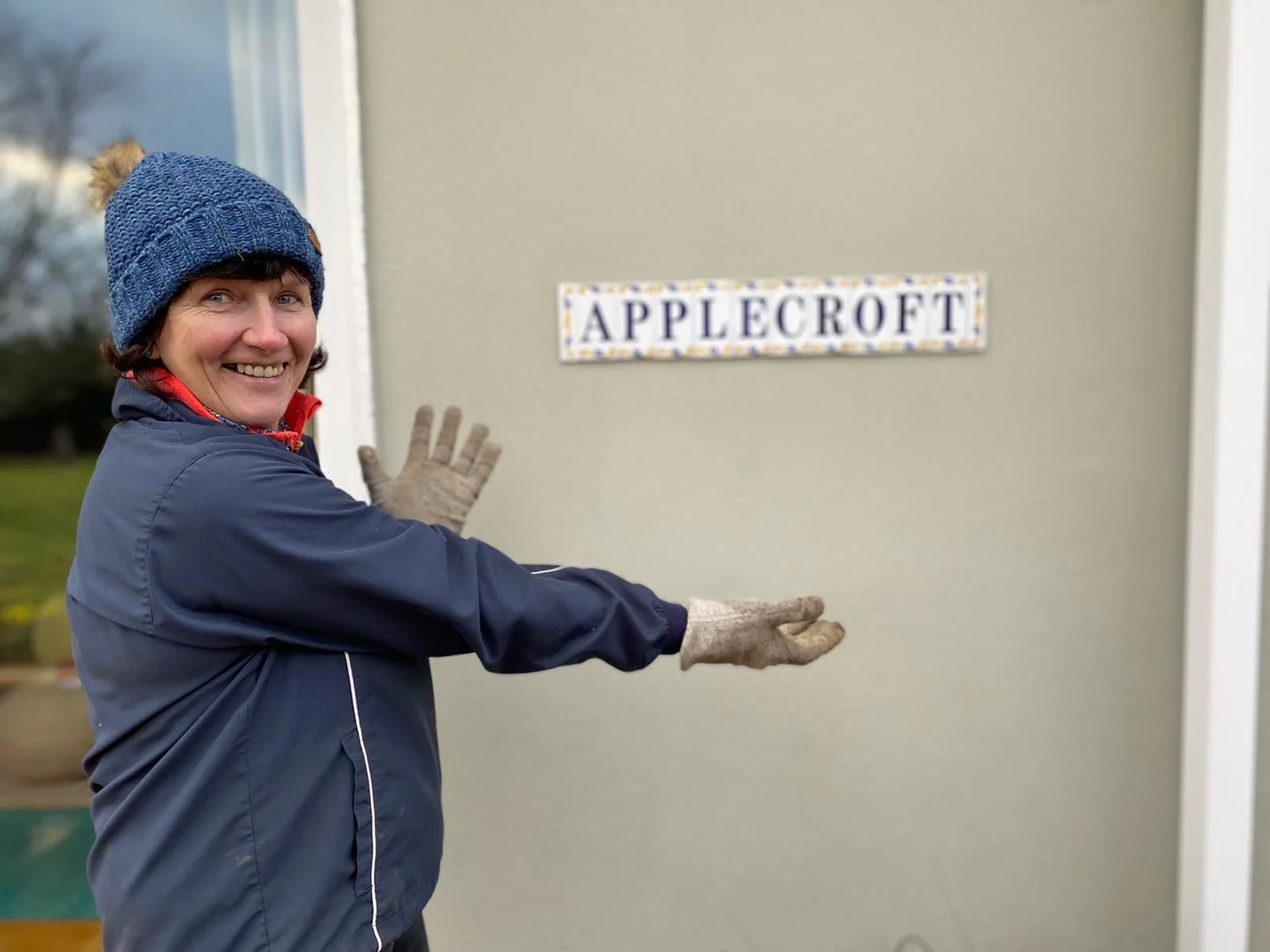 Woman in a blue jacket and knit hat standing outside and pointing at a decorative sign that says "APPLECROFT."