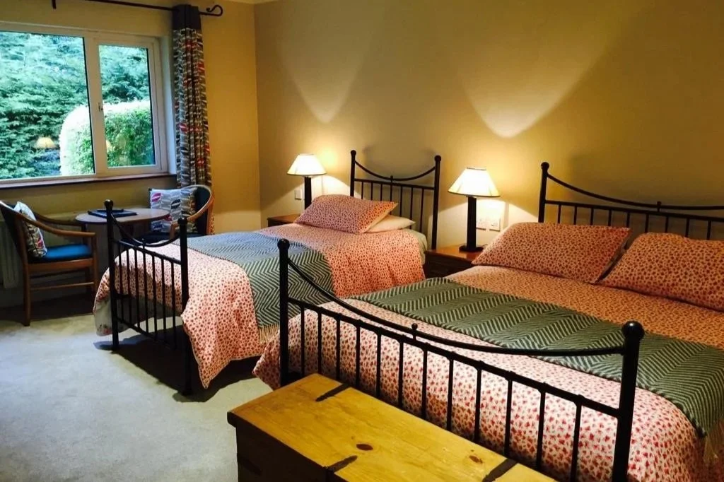 A bedroom with two single beds and one large bed, all with matching pink and red patterned bedding, situated against a yellow wall. There are two bedside lamps between the beds, a window with a view of greenery, and a small seating area with a table 