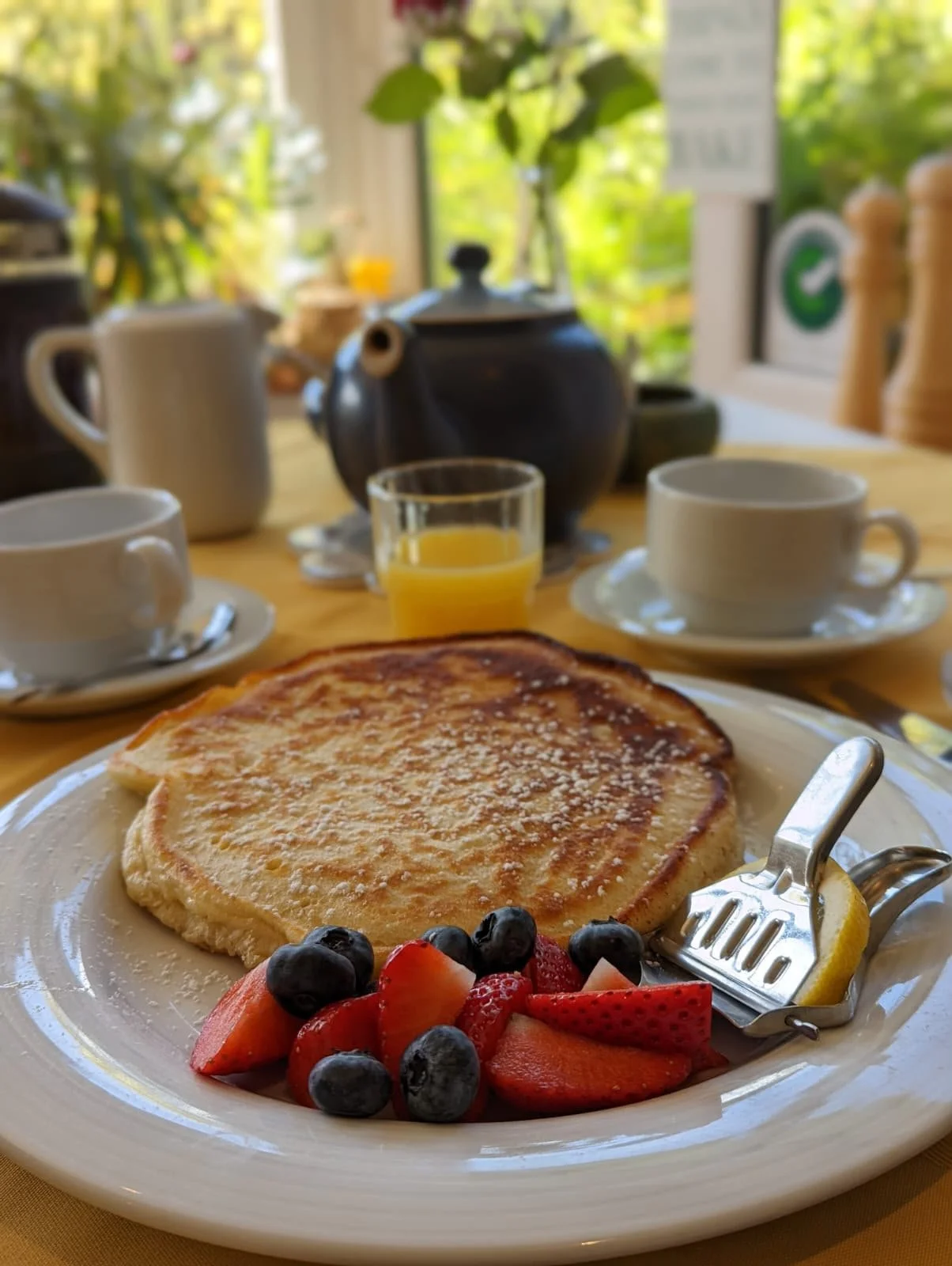 A breakfast plate with a stack of pancakes topped with powdered sugar, alongside fresh fruit including strawberries, blueberries, and a wedge of lemon. In the background, there are cups of coffee, a glass of orange juice, and a teapot on a table near a window with greenery outside.