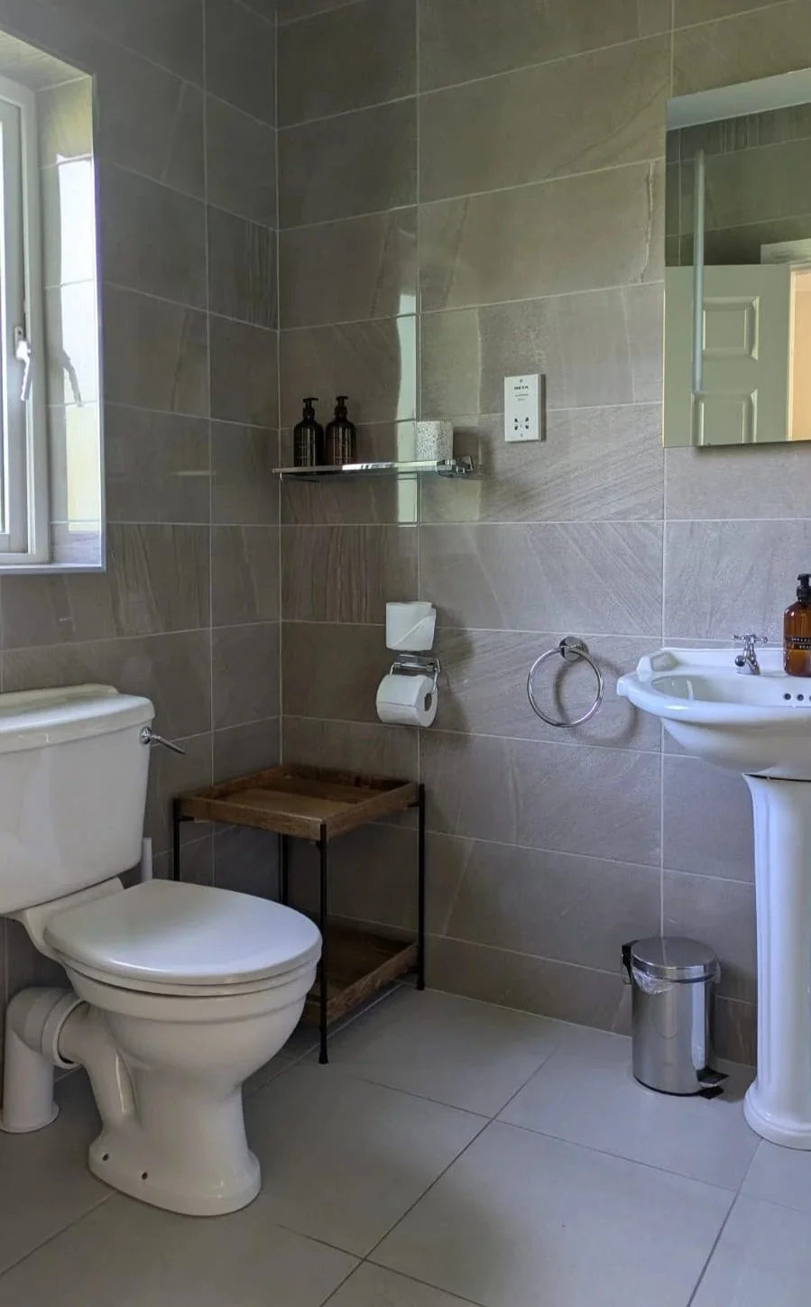 A modern bathroom with beige tiled walls and floor, a white toilet, a white pedestal sink with a bottle of hand soap, a small mirror, a wall-mounted toilet paper holder, a metal trash can, a small wooden and metal shelf, and a window allowing natural